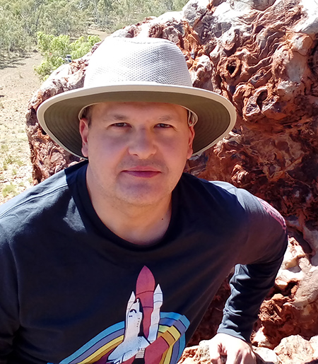 Man in hat with rainbow space shuttle design on t-shirt, with boulder behind him.