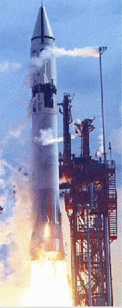 Rocket launch, flame and smoke below a stocky white rocket rising next to a launch tower.
