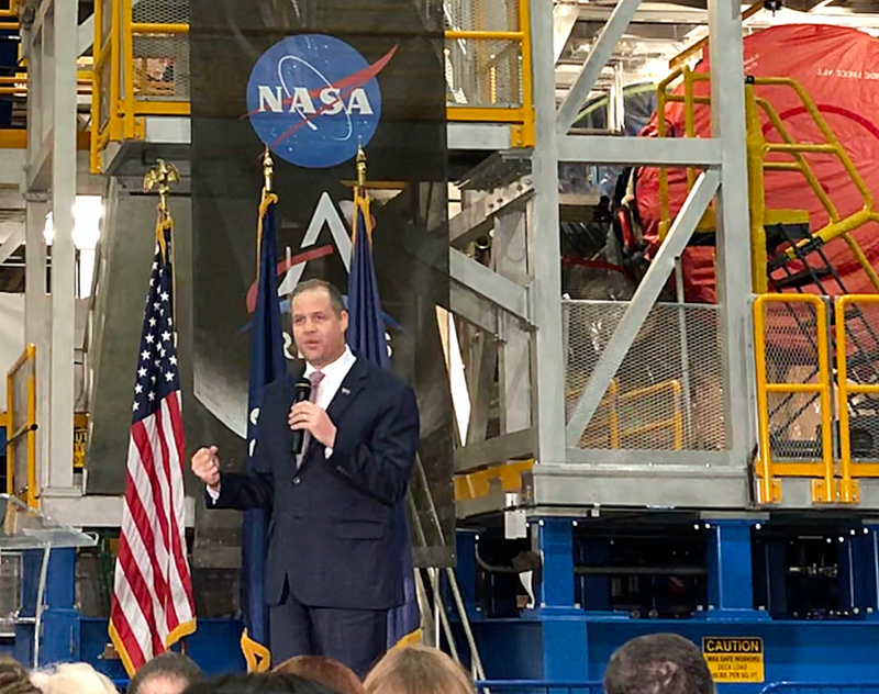 A man in a suit standing under a NASA logo in front of a machine assembly.