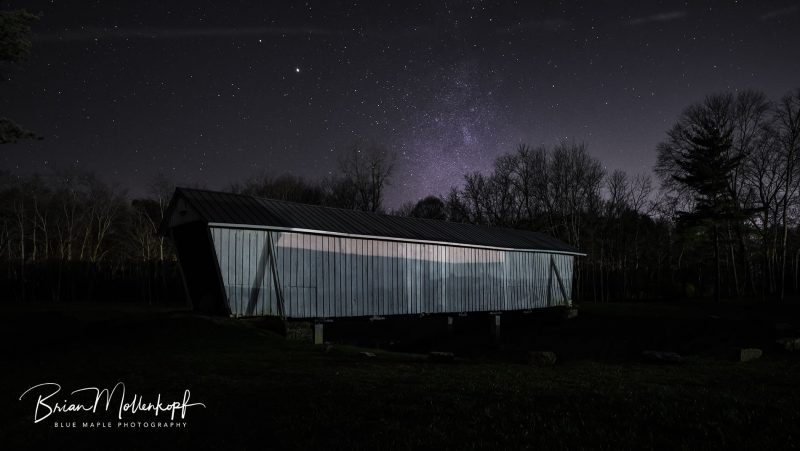 Saturn and Jupiter float beside the Milky Way over an antique wooden covered bridge.