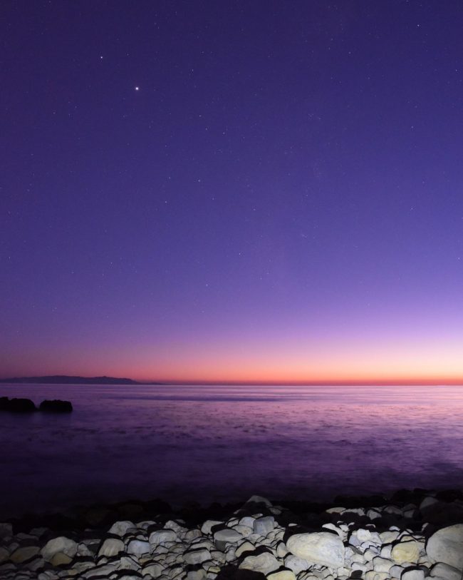 Jupiter and Saturn above sunset over calm water with small rocks in foreground.