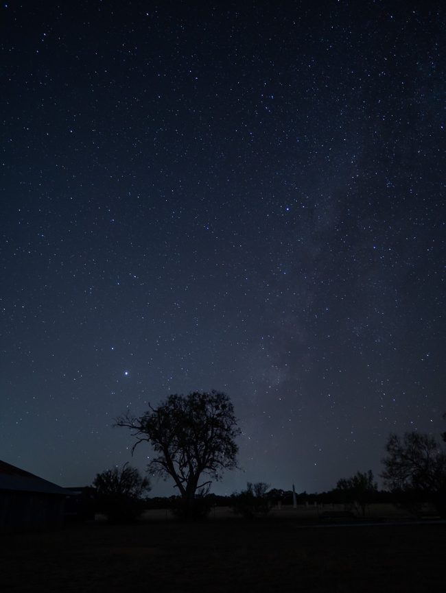 Jupiter, Saturn, Milky Way in starry sky over dark landscape with trees and barn.