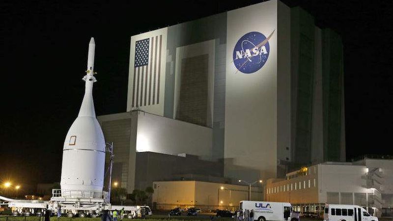 Night view of stocky white rocket near gigantic building with flag and NASA logo on it.