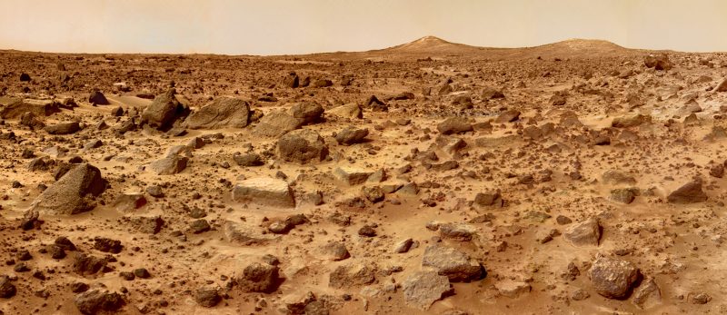 Reddish rocky terrain with two hills in the distance and dusty sky.