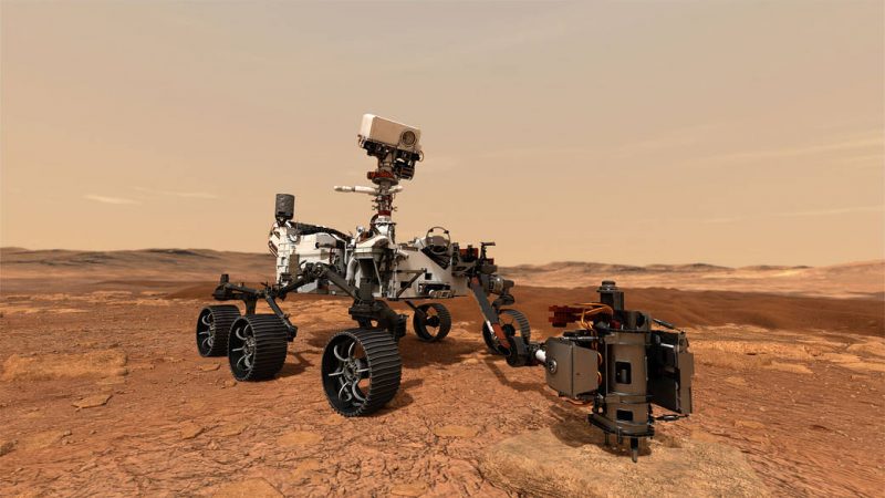Robotic rover with six wheels on brownish-reddish terrain, with hills in distance and dusty sky above.