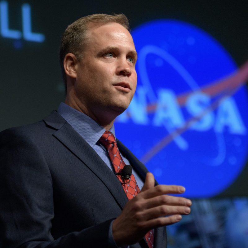 A man in a suit, next to a large NASA logo.