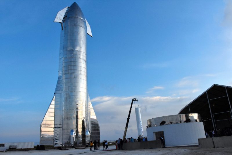 A tall metallic rocket stands on its fins, towering over onlookers, with a clear sky backdrop.