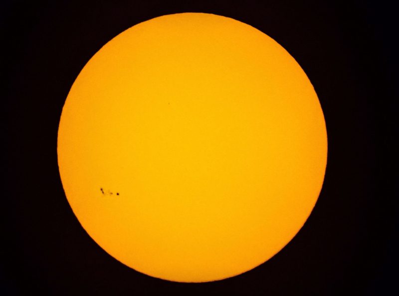 Round yellow globe of sun, with small set of spots and blotches nearing the left limb, in about the 8 o'clock position.