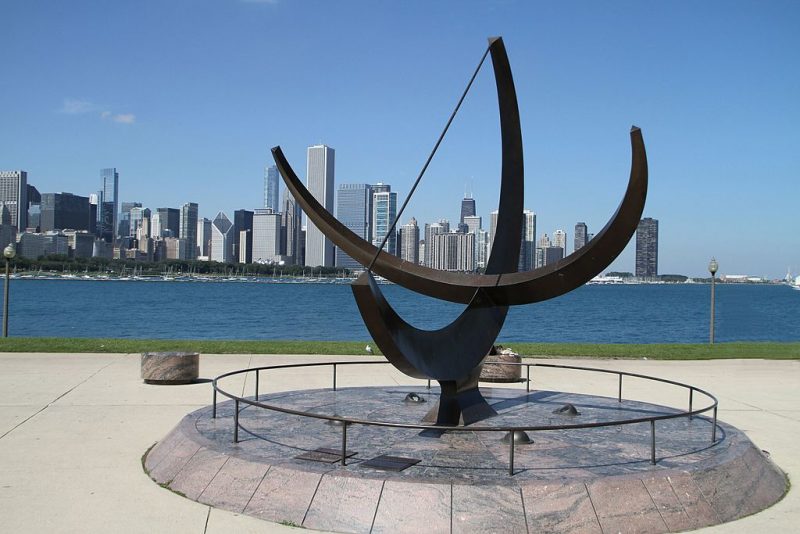 Giant sundial made of 2 intersecting semicircular bars, with city skyline beyond.