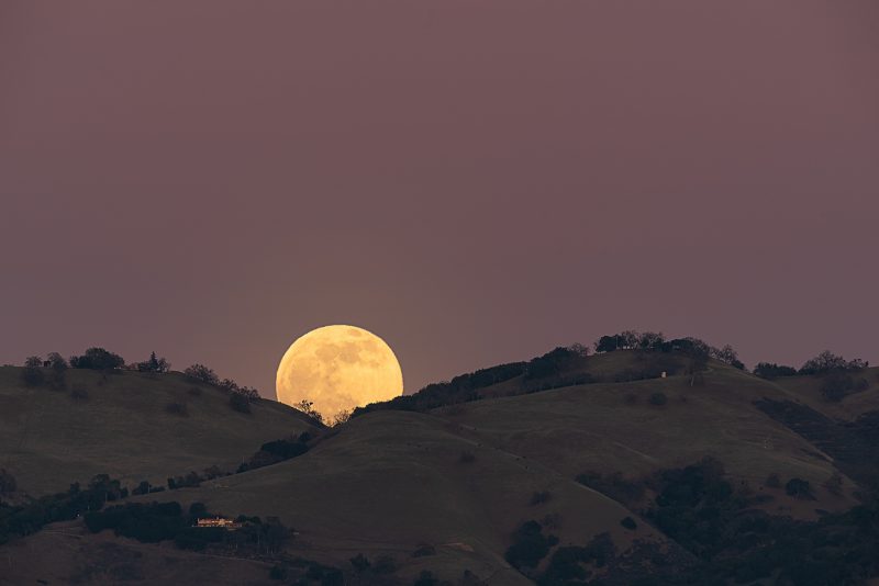 Full yellow moon peeps out from valley between two scrub-covered hills.