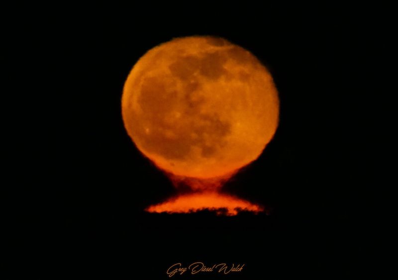 Orange moon against dark sky with a sliver of reflection below it.