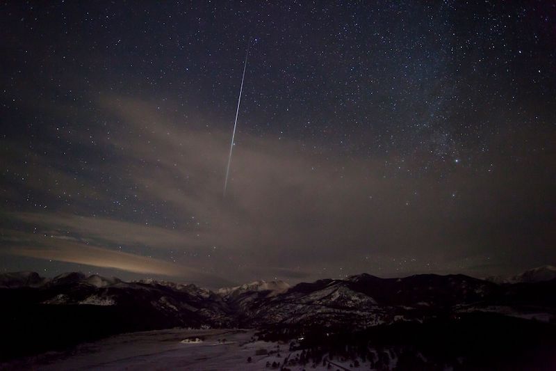 Nearly vertical, bright, thin white streak through light clouds over dark mountains.