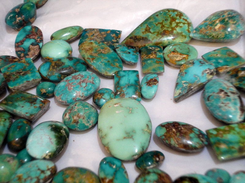 About 40 pieces of the December birthstone turquoise, all polished, mostly oval but a few rectangular, scattered on a white surface. Each piece has different shades of green with brown veining.