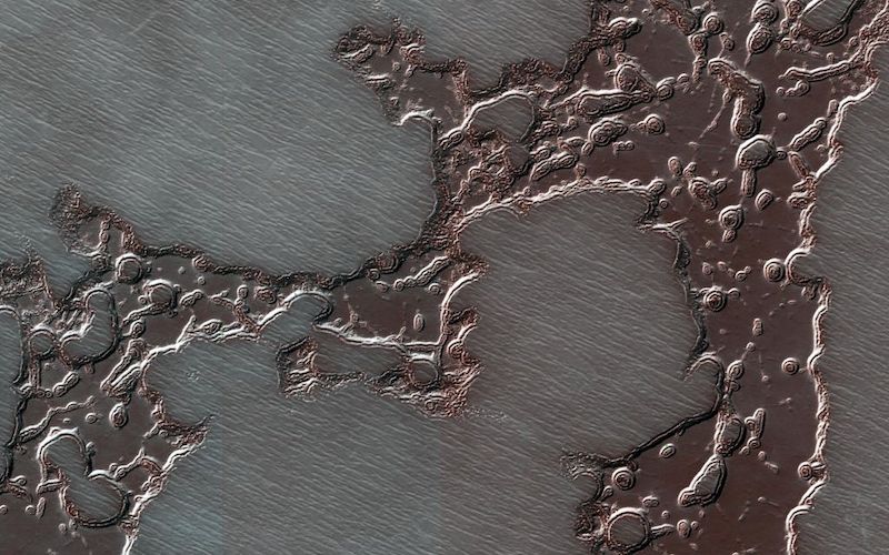 Dark reddish splotch-like patches of frozen water ice are pictured on a striated surface.