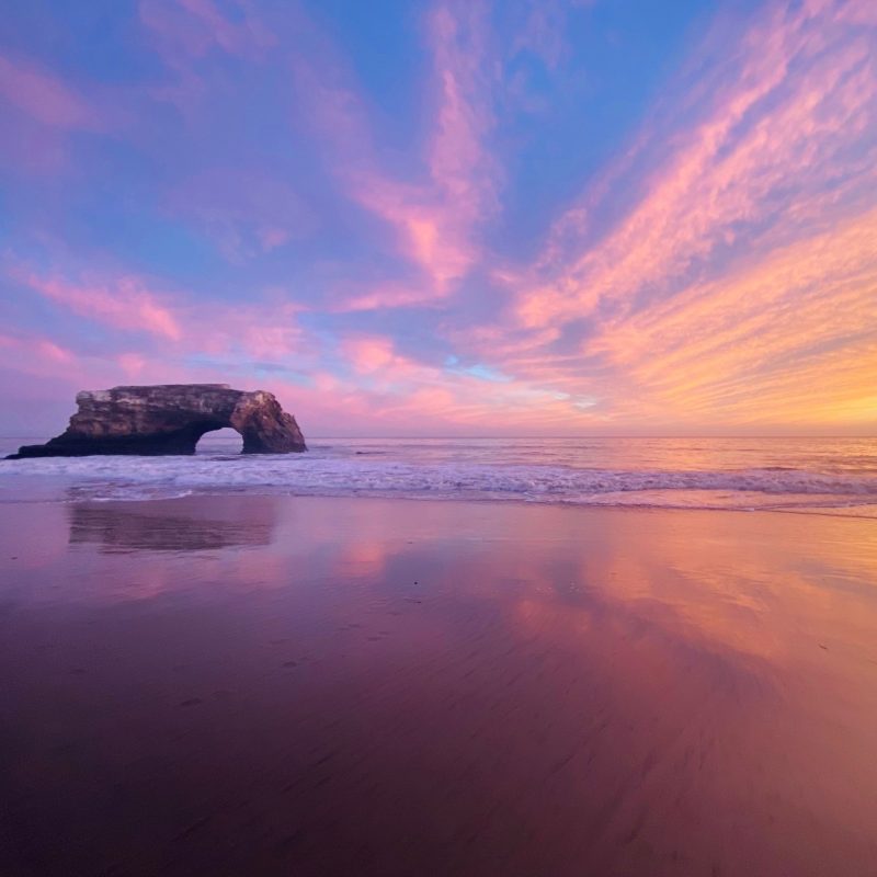A beach at sunset, a stone arch on one side and colorful pink streaky clouds in a soft blue sky.