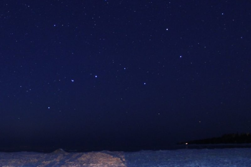 Star field with prominent Big Dipper stars in deep blue sky over icy lake.