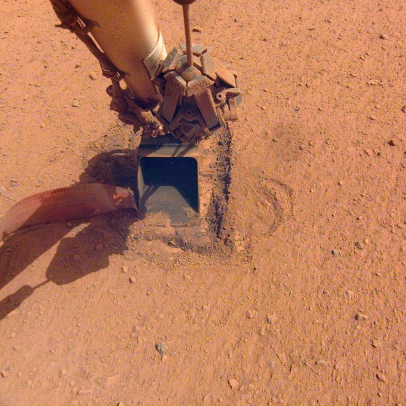 Long metallic cylindrical arm with shovel at the end, on reddish ground.