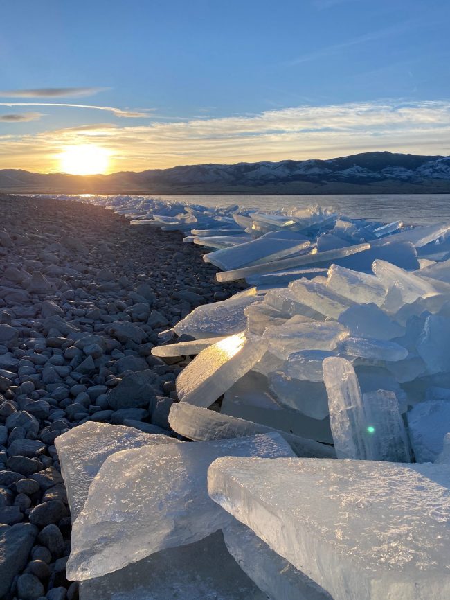 Slabs of ice piled up on lakeshore with lowering sun glinting off them.