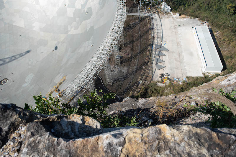 Edge of parabolic structure and 5 11-meter gray triangles lying on the ground beside it looking tiny.