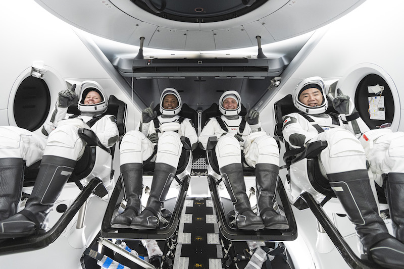 Four men wearing white SpaceX spacesuits, strapped into their seats and ready for launch.