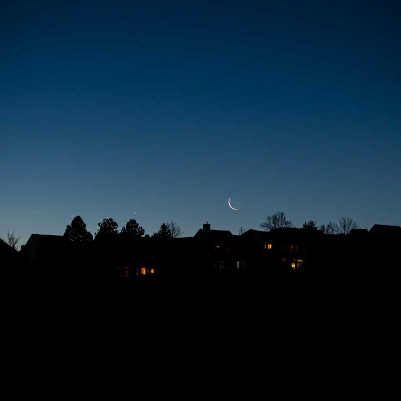 Venus and thin crescent moon in deep blue sky near dark line of silhouetted rooftops.