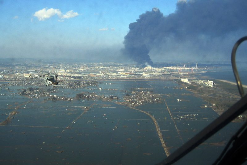 Aerial view of seaside port inundated by water, huge column of black smoke in distance.