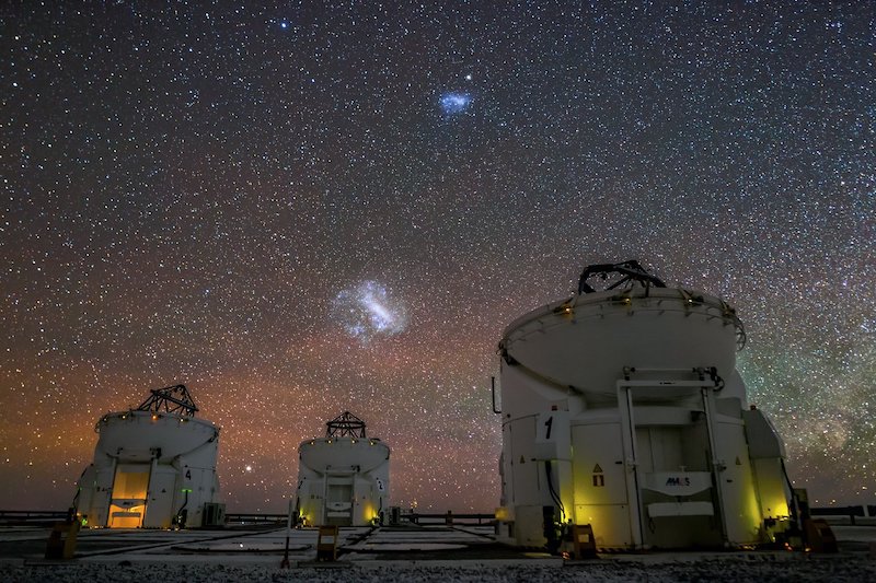 Two irregular glowing fuzzy patches on the sky over several large telescope domes. 