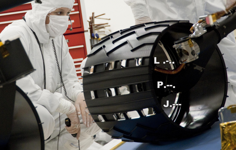 Large, wide black wheel with pattern of holes in it and man in white protective lab suit.