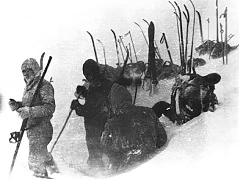 Black-and-white photo of skiers breaking snow, with their skis stashed upright behind them.