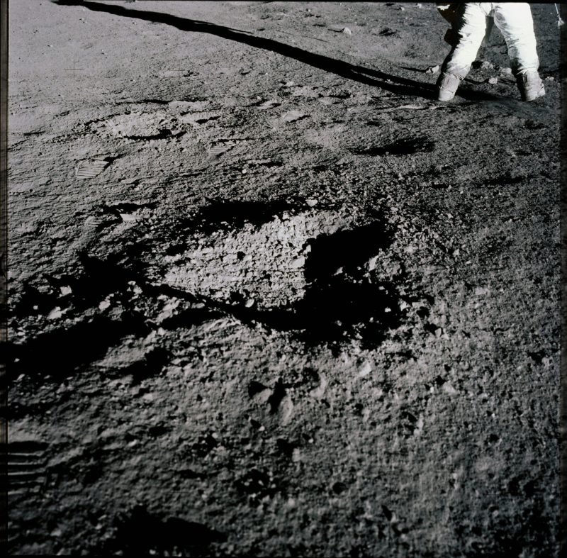 Rocky gray ground with a shallow hole the shape of a heart, space-suited legs.