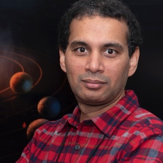 Smiling man in checkered shirt, with two planets behind him.