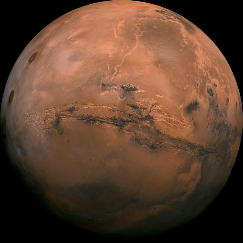 A nearly full reddish Mars seen from orbit, with a giant dark slash covering near a whole hemisphere.