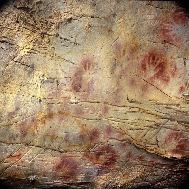 Handprints surrounded by red, like a stencil, on a cracked, brown rock wall.