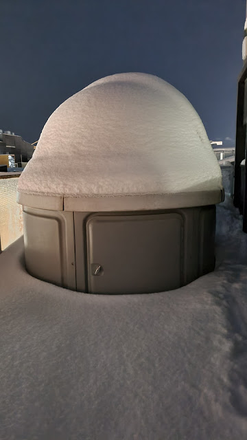 Heavily snowcapped observatory in the shape of a domed beige cylinder surrounded by deep snow.