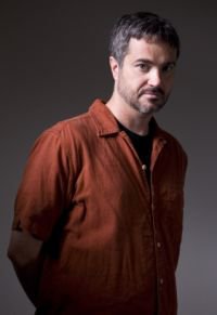 Short-haired bearded man in red shirt on plain grayish background.