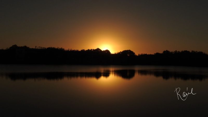 Sun barely peeking over forested horizon under bronze sky, reflected in foreground lake.