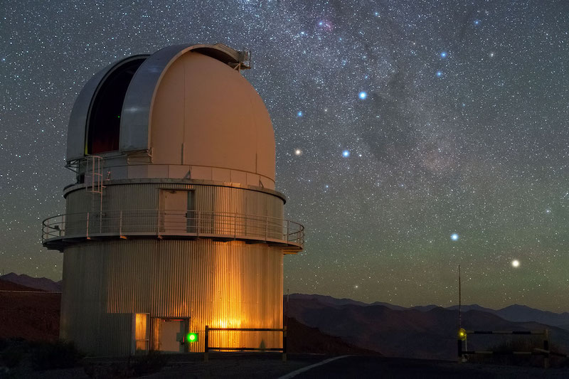 Dense star field with Milky Way and several bright stars behind a domed observatory building.