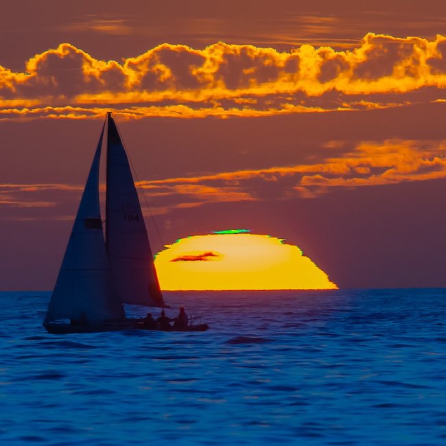 Sailboat in solhouette in front of a setting sun. The sun has a top layer of green.
