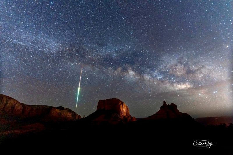 Red rock formations with fuzzy band of stars and vertical streak of light.
