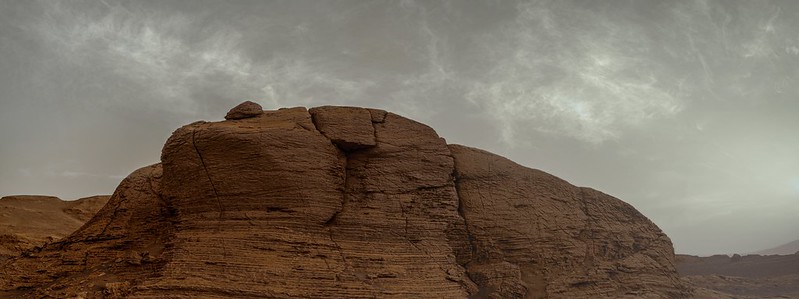 Reddish, rounded, rock outcrop with rock bump on top and white wispy clouds in sky above.