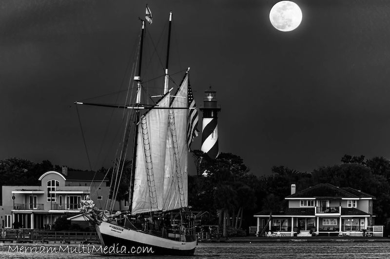 Stark grayscale of sailboat, lighthouse, seaside houses, and a full moon at top right. 