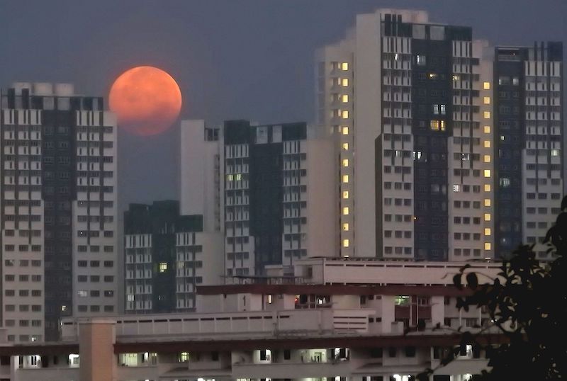 Orange orb, darker on the bottom, peeking out from between high-rise buildings in deep twilight.