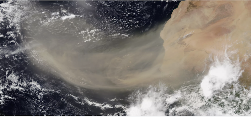 Orbital view of Earth with wide tan streak reaching from Africa out across the cloud-spotted blue Atlantic ocean.