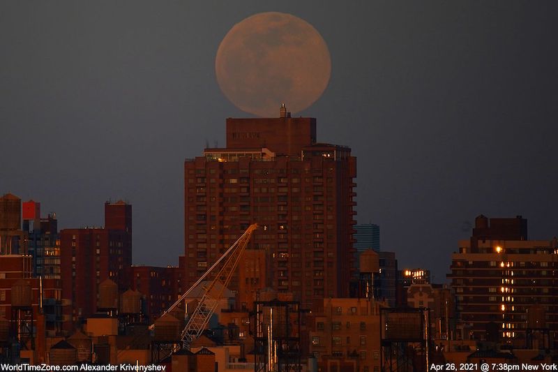 Large and faint full moon seemingly resting on top of a reddish high rise building.