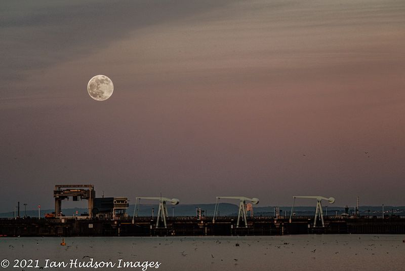 Detailed full moon on wispy pink sky above drawbridges, with water in the foreground.