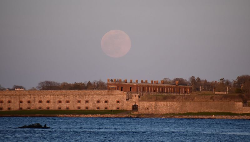 Faint pink full moon over old stone fort with water in the foreground.