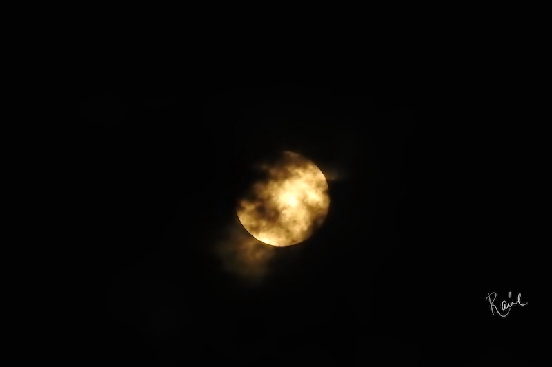 Bright yellow moon shining through patchy, obscuring black clouds on black background. 