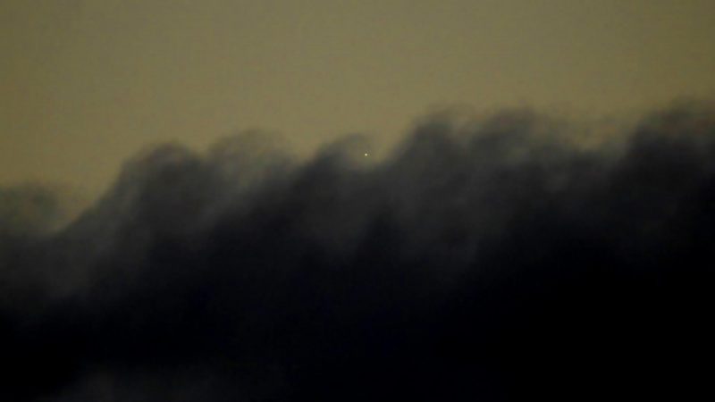 Tiny bright dot in dim sky just above wave-shaped dark gray clouds.