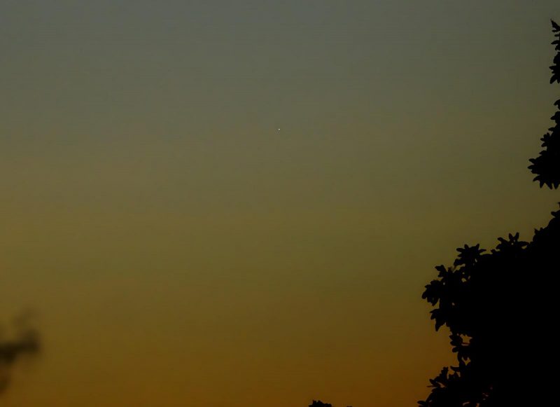 A faint dot of light in a dim deep orange to blue twilight sky, with trees in the foreground.