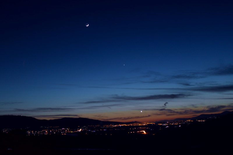 A colorful sunset with a thin crescent moon, overlooking a small city nestled in a valley, together with two bright planets.